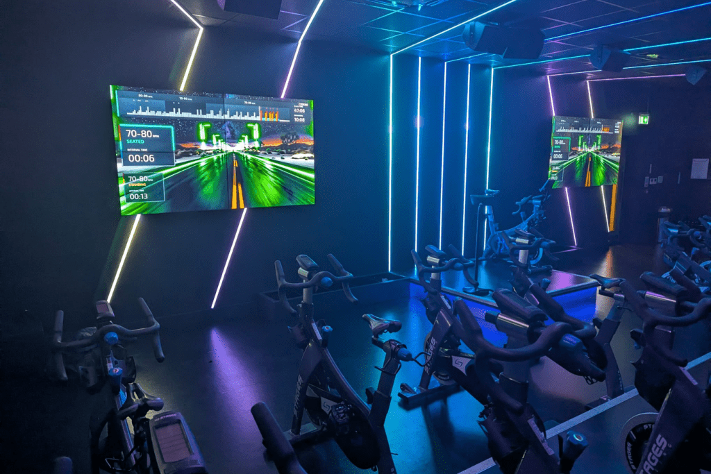 A neon-lit indoor cycling studio where the existing bike fleet is already in place, with CicloStudio class output — a virtual ride, interval profile and cadence targets — running on two wall-mounted screens.