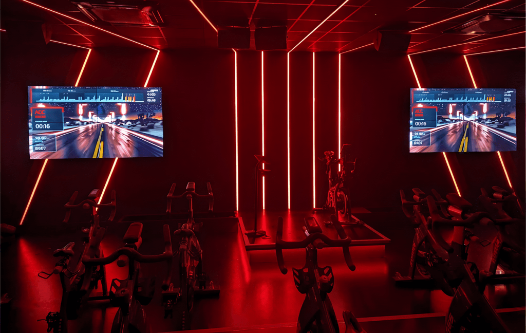 An indoor cycling studio lit in red, fully equipped with bikes and two studio screens running a virtual ride, with no riders in the room.